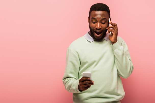Amazed african american man talking on smartphone and using cellphone on pink background  - Fotoğraf, Görsel