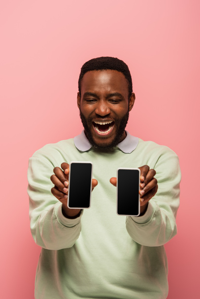 Hombre afroamericano positivo sosteniendo teléfonos inteligentes en primer plano borroso aislado en rosa  - Foto, Imagen