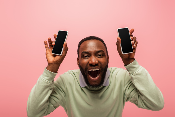 Emocionado hombre afroamericano con teléfonos móviles mirando a la cámara aislada en rosa  - Foto, imagen