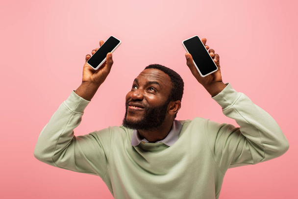 Смартфони в руках усміхненого афроамериканського чоловіка, ізольованого на рожевому  - Фото, зображення