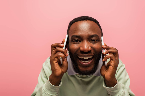 Uśmiechnięty Afroamerykanin rozmawiający na smartfonach odizolowanych na różowo  - Zdjęcie, obraz