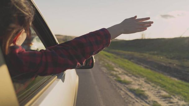 Το κορίτσι ταξιδεύει με το χέρι της έξω από το παράθυρο του αυτοκινήτου και να πιάσει τον άνεμο στο φως του ήλιου, λαμβάνοντας ένα μακρύ δρόμο σε ένα επαγγελματικό ταξίδι, ένας ευτυχισμένος άνθρωπος παίζει με τον άνεμο, το χρόνο του να ονειρευτεί ελεύθερα - Φωτογραφία, εικόνα