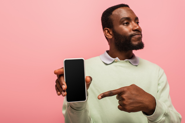Hombre afroamericano borroso apuntando a teléfono inteligente con pantalla en blanco aislado en rosa  - Foto, imagen
