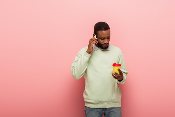 African american man with coffee to go talking on mobile phone on pink background  - Photo, Image