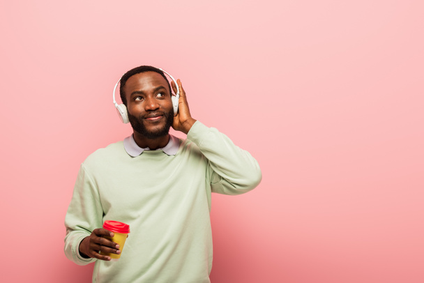 Αφροαμερικάνος με ακουστικά κρατώντας χάρτινο κύπελλο σε ροζ φόντο  - Φωτογραφία, εικόνα