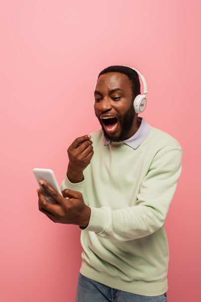 Amazed african american man in headphones using mobile phone isolated on pink  - Foto, afbeelding