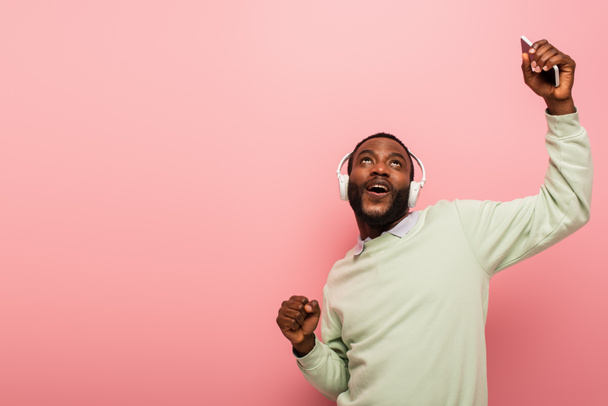 Homme afro-américain dans un casque montrant un geste oui et tenant le téléphone portable isolé sur rose  - Photo, image