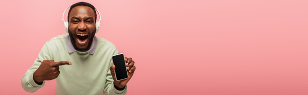 アフリカ系アメリカ人の男性ヘッドフォンで指差すピンク、バナーに隔離された空白の画面を持つスマートフォン  - 写真・画像