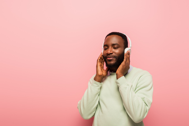 Αφροαμερικάνος με ακουστικά που ακούει μουσική με κλειστά μάτια σε ροζ φόντο  - Φωτογραφία, εικόνα