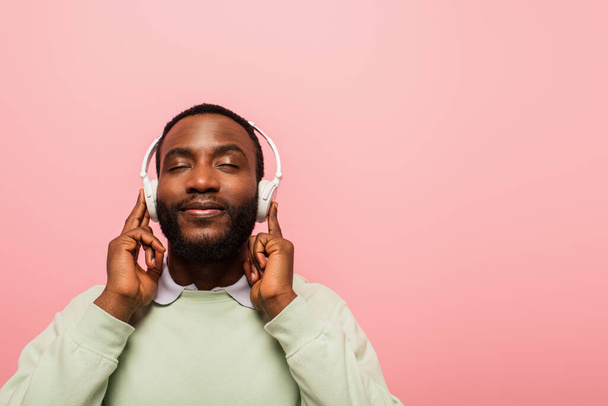 Jeune homme afro-américain utilisant des écouteurs sans fil isolés sur rose  - Photo, image