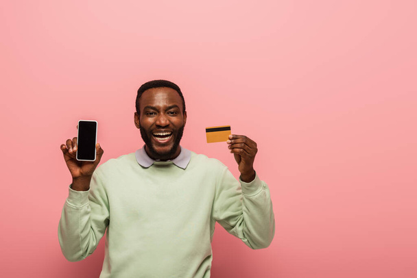 Fröhlicher afrikanisch-amerikanischer Mann mit Handy mit leerem Bildschirm und Kreditkarte auf rosa Hintergrund  - Foto, Bild