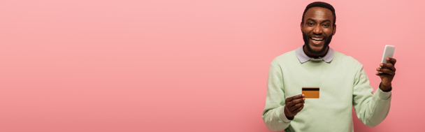 Щасливий афроамериканець, який користується кредитною карткою і смартфоном, ізольований на рожевому банері.  - Фото, зображення