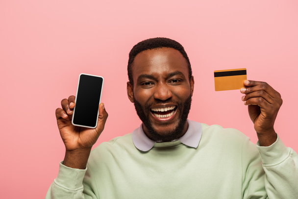 Hombre afroamericano positivo sosteniendo teléfono inteligente y tarjeta de crédito aislado en rosa  - Foto, Imagen
