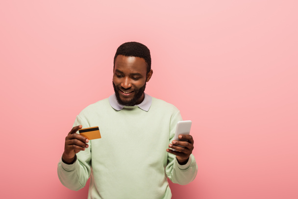 African american man using smartphone and credit card on pink background  - Фото, изображение
