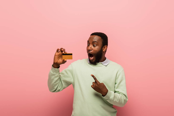 Aufgeregter afrikanisch-amerikanischer Mann zeigt auf Kreditkarte auf rosa Hintergrund  - Foto, Bild