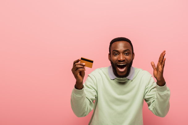 erstaunt afrikanisch-amerikanischer Mann schaut in die Kamera, während er Kreditkarte isoliert auf rosa zeigt - Foto, Bild