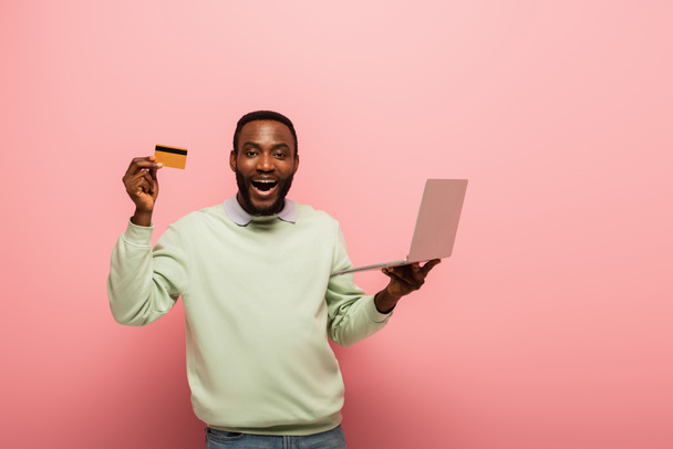ピンクの背景にノートパソコンを持ちながらクレジットカードを見せるアフリカ系アメリカ人男性に驚きました - 写真・画像