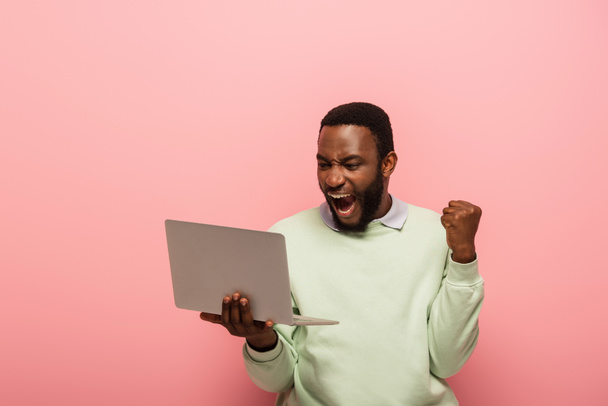 ενθουσιασμένοι Αφροαμερικανός άνδρας με φορητό υπολογιστή δείχνει κερδίσει χειρονομία και φωνάζοντας απομονώνονται σε ροζ - Φωτογραφία, εικόνα