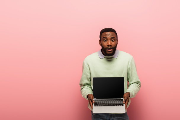 sorprendido hombre afroamericano mostrando portátil con pantalla en blanco sobre fondo rosa - Foto, imagen