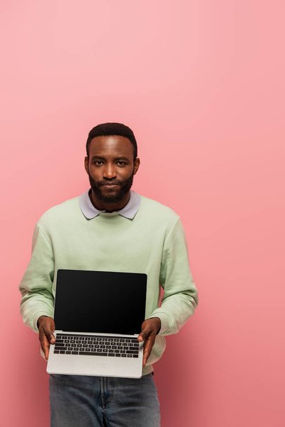 african american man looking at camera while holding laptop with blank screen on pink background - Fotografie, Obrázek