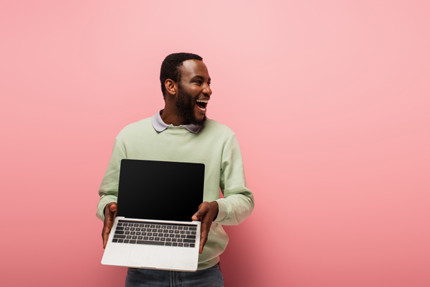 hämmästynyt afrikkalainen amerikkalainen mies katselee pois ja pitää kannettava tietokone tyhjä näyttö vaaleanpunainen tausta - Valokuva, kuva