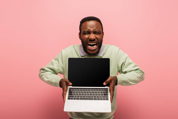 hombre afroamericano emocionado con la boca abierta que muestra el ordenador portátil con la pantalla en blanco en rosa  - Foto, imagen
