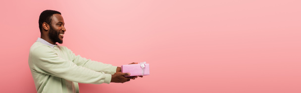 uśmiechnięty Afrykanin amerykański mężczyzna trzymając pudełko prezentów w wyciągniętych rękach odizolowanych na różowy, sztandar - Zdjęcie, obraz