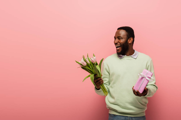 laughing african american man with gift box and tulips looking away on pink background - Φωτογραφία, εικόνα
