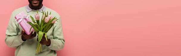 rajattu näkymä hymyilevä afrikkalainen amerikkalainen mies lahjapaketti ja tulppaanit eristetty vaaleanpunainen, banneri - Valokuva, kuva
