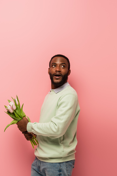 asombrado hombre afroamericano mirando hacia otro lado mientras sostiene tulipanes sobre fondo rosa - Foto, imagen