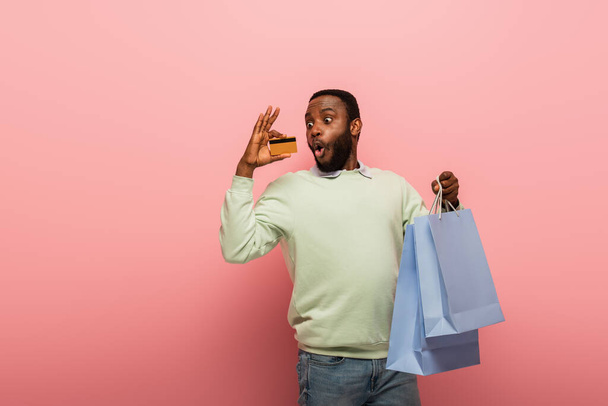 sorprendido hombre afroamericano mirando a la tarjeta de crédito mientras que la celebración de compras en fondo rosa - Foto, Imagen