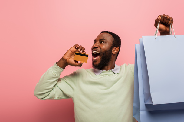 excited african american man looking away while holding credit card and purchases on pink background - Photo, Image
