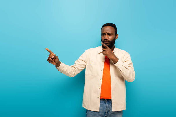 thoughtful african american man touching face while pointing aside with finger on blue background - Photo, Image