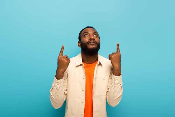 young african american man looking up and pointing with fingers isolated on blue - Foto, afbeelding