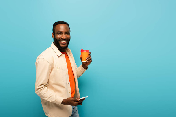 smiling african american man holding takeaway drink while using cellphone isolated on blue - Fotoğraf, Görsel