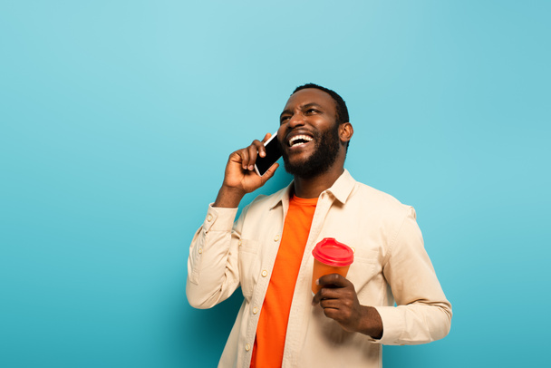 smějící se africký Američan muž s odnést drink mluvit na smartphone na modrém pozadí - Fotografie, Obrázek