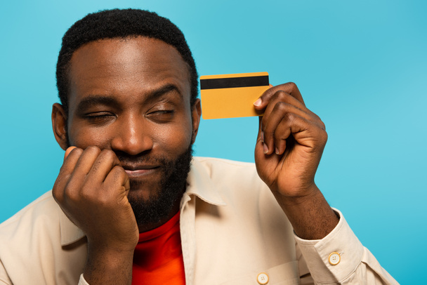 positive african american man dreaming while holding credit card isolated on blue - Photo, Image