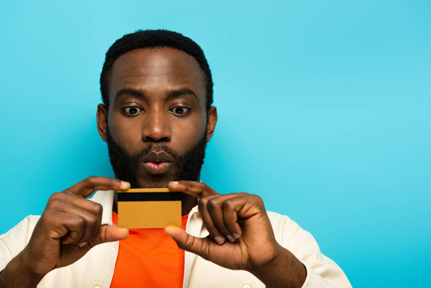 verrast Afrikaans amerikaanse man tonen credit card geïsoleerd op blauw - Foto, afbeelding