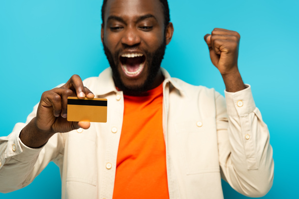 izgatott afro-amerikai férfi kiabálva és mutatja hitelkártya elszigetelt kék, homályos háttér - Fotó, kép