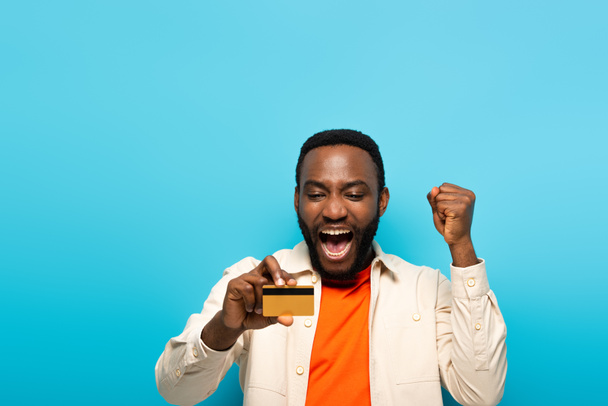 hombre afroamericano emocionado con tarjeta de crédito gritando y mostrando gesto de regocijo aislado en azul - Foto, Imagen