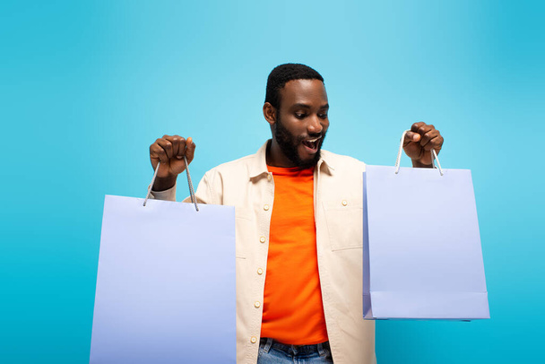 amazed african american man holding shopping bags isolated on blue - Fotoğraf, Görsel