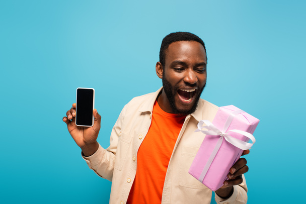 erstaunt afrikanisch-amerikanischer Mann mit Geschenkbox und Smartphone mit leerem Bildschirm isoliert auf blau - Foto, Bild