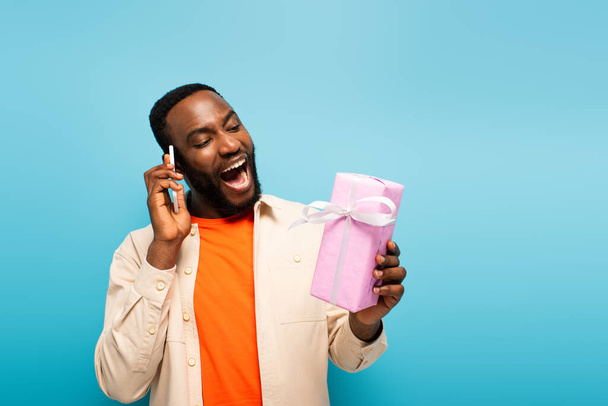 espantado homem americano africano olhando para o presente enquanto fala no smartphone isolado no azul - Foto, Imagem