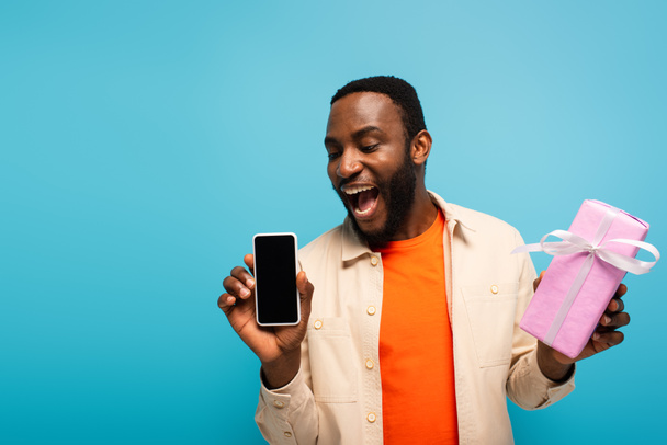 amazed african american man showing smartphone and gift box isolated on blue - 写真・画像