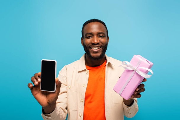 χαμογελαστός Αφροαμερικάνος που κρατά το παρόν και το smartphone απομονωμένο στο μπλε - Φωτογραφία, εικόνα