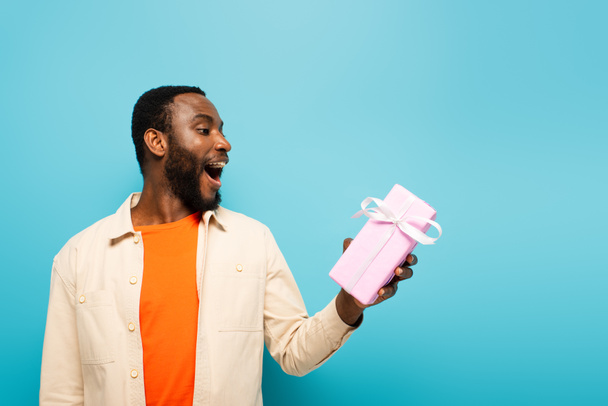 amazed african american man holding present isolated on blue - 写真・画像