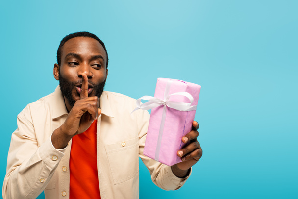 joven afroamericano hombre mostrando signo secreto mientras sostiene caja de regalo aislado en rosa - Foto, Imagen