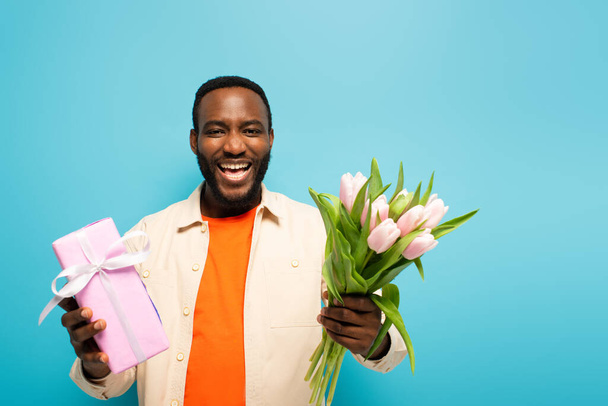 gelukkig Afrikaans amerikaanse man met geschenk doos en tulpen lachen op camera geïsoleerd op blauw - Foto, afbeelding