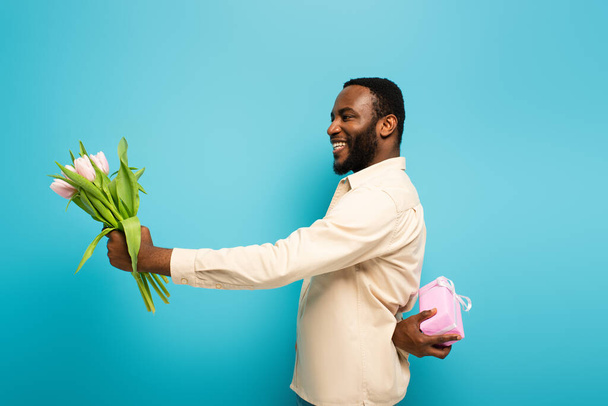 radostný africký Američan představující tulipány při držení dárkové krabice za zády na modrém pozadí - Fotografie, Obrázek