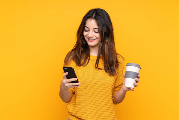 Jonge blanke vrouw geïsoleerd op gele achtergrond met koffie mee te nemen en een mobiele - Foto, afbeelding